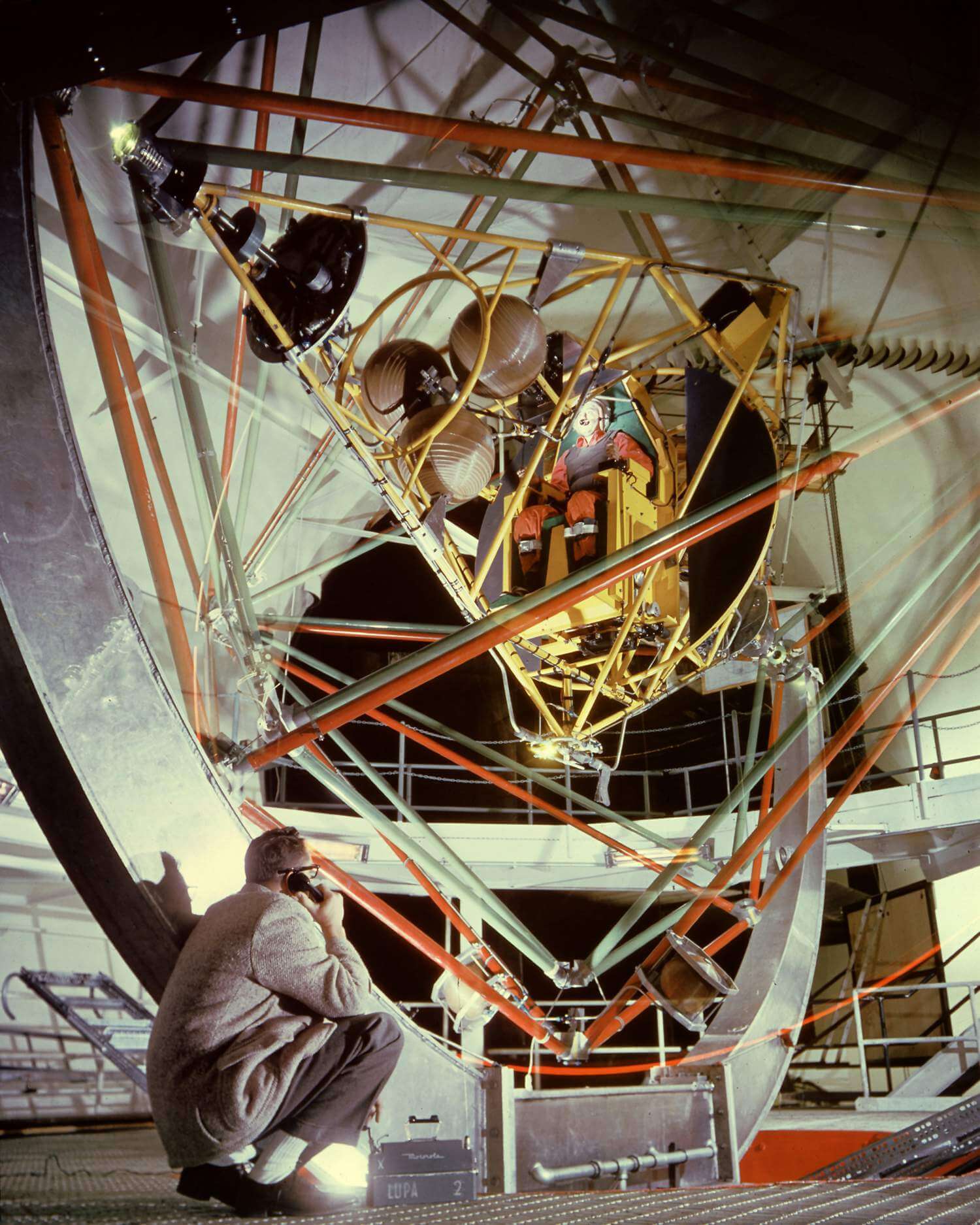 An archival photograph of a multi-axis gimbal test rig, with yellow machinery, rust-orange structural rails, and sage-green metalwork.